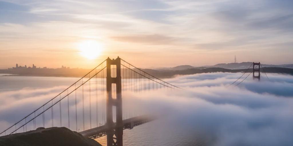Die Golden Gate Bridge steigt bei Sonnenaufgang aus tiefem Nebel empor, die Pfeiler in International Orange vor blassem Himmel